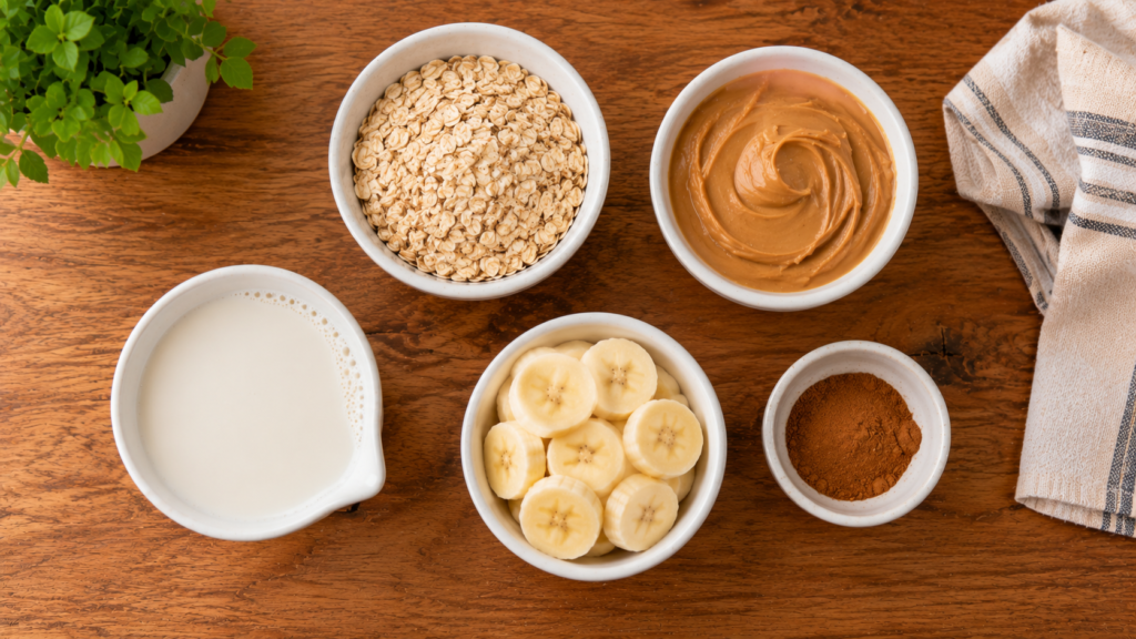 Ingredients for peanut butter banana overnight oats including rolled oats, peanut butter, banana, plant-based milk, and cinnamon arranged on a wooden countertop.