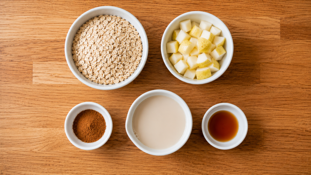 Ingredients for a cinnamon pear oat bowl including rolled oats, diced pears, cinnamon, plant-based milk, and vanilla arranged on a wooden countertop.